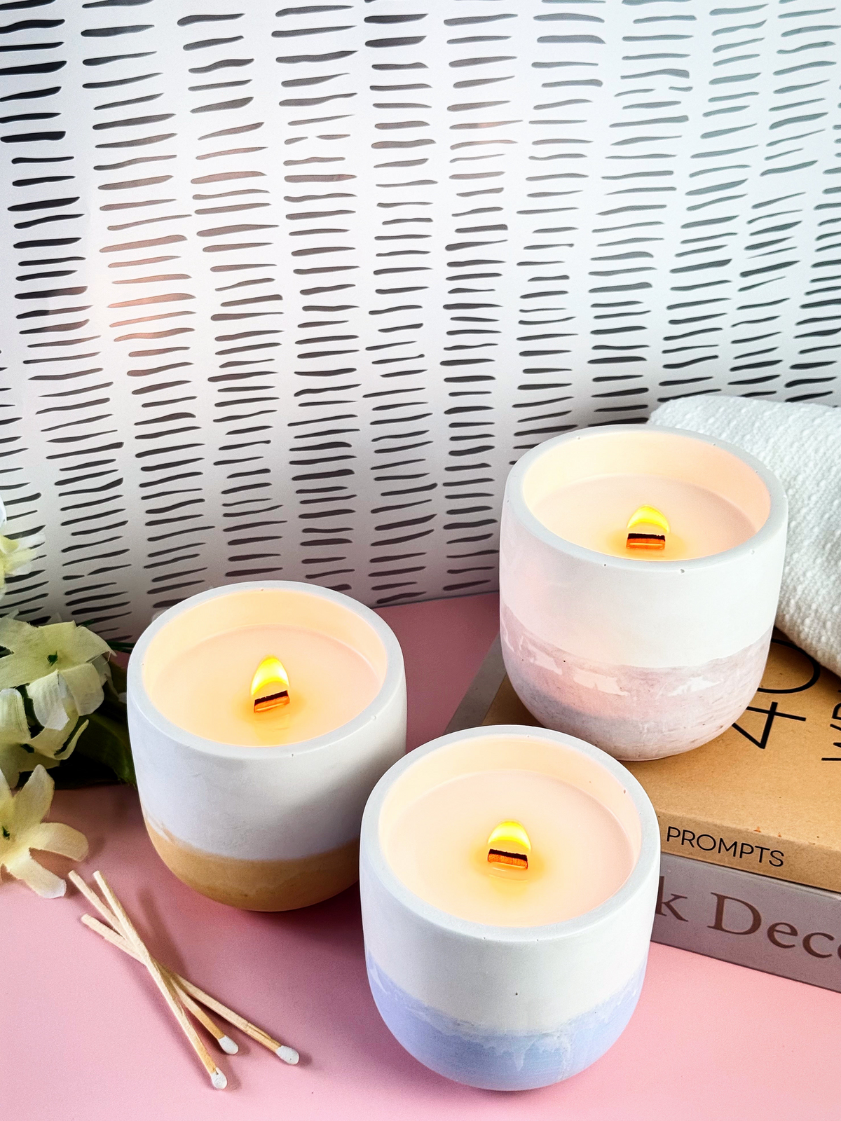 Three lit wax candles in concrete holders with white, beige, and pastel blue color-blocked designs, arranged on a pink surface with a woven backdrop and decorative props.